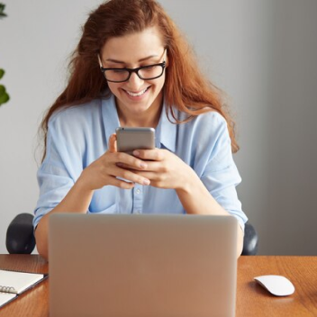 Mulher ruiva com óculos segura o celular e sorri enquanto trabalha em frente a um notebook em ambiente interno.
